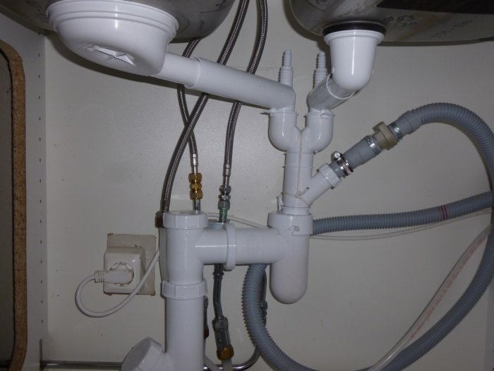 Installation under diskbänk inkluderar vita avloppsrör, slangar och anslutningar mot grått väggunderlag.