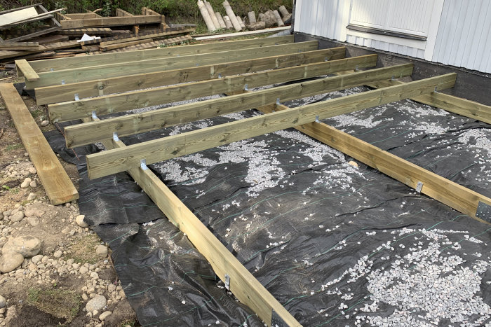 Grundläggningsarbete för byggprojekt med träbärlinor ovanpå markduk och spridda makadamstenar.