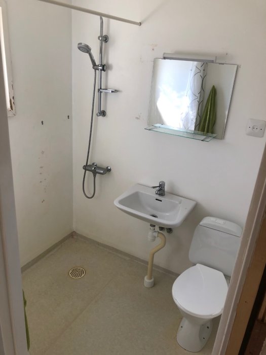 Äldre badrum i behov av renovering med vitkaklad dusch, handfat och toalett mot en bakgrund av släta väggar.