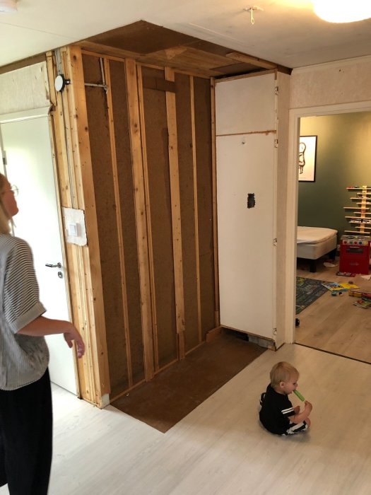 Interiör av ett hem under renovering med synlig träregelvägg och ett barn på golvet.