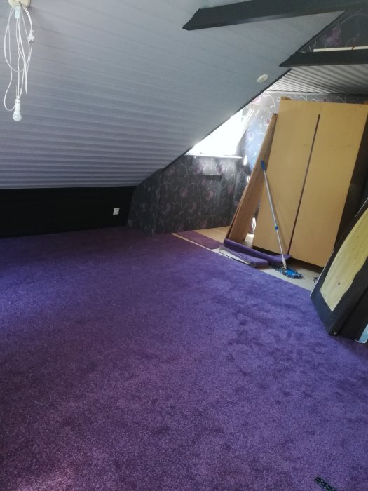 Inredningsbild av ett vindsrum med lila matta, lutande tak, otydlig väggstruktur utan synliga reglar och brädor lutade mot väggen.