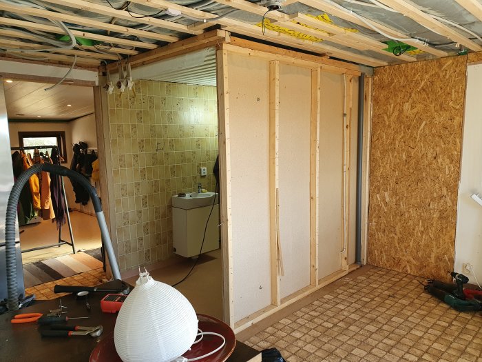 Renoveringsarbete i ett rum med öppet tak och väggar före isolering, gammal handfat och verktyg synliga.