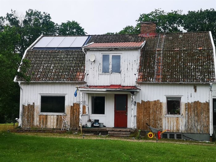 En vit, sliten villa med mansardtak, behövande renovering och solpaneler på taket.