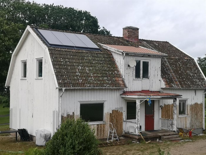 Renoveringsobjekt med slitet mansardtak, solarpaneler och flagnande vit fasad på hus i behov av omvårdnad.