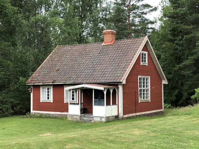 Rödmålat äldre hus med slitet tegeltak, byggt på 1920-talet, omgivet av gröna träd och gräs.