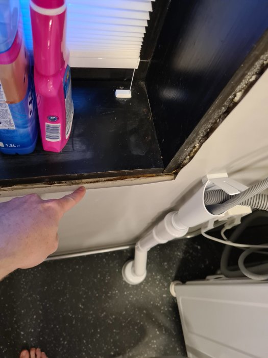 Hand pekar på outredda lister och exponerad gipsskiva vid fönster i ett pågående badrumsrenoveringsprojekt.