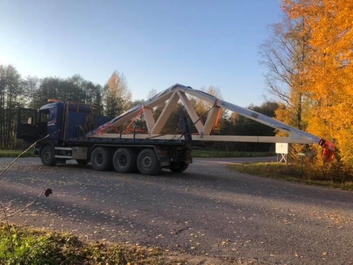 Lastbil som transporterar en stor takkonstruktion på en väg omgiven av höstlöv.
