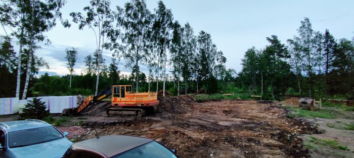 Grävmaskin på en byggarbetsplats med förberudd grund för gjutning omgärdat av träd i skymningen.