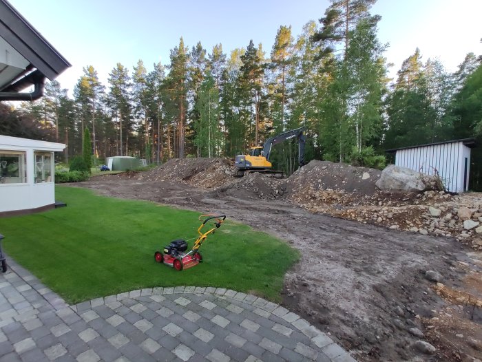 En gräsmatta framför ett hus med en gräsklippare, grävningsarbete och en grävmaskin i bakgrunden på en lerig mark.