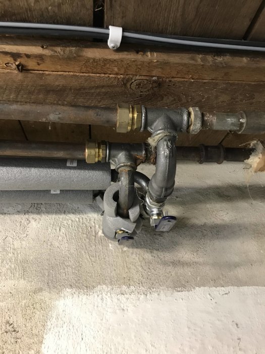 Värmerör med T-koppling i källare, isolerade rör från värmesystem med etiketter.
