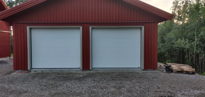 Dubbla garageportar på en röd träbyggnad med vit foder, grusbana framför och skog i bakgrunden.