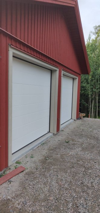 Nyinstallerade vit garageportar inramade med träpanel på en rödmålad byggnad med tidigt skede av landskapsarbete i förgrunden.