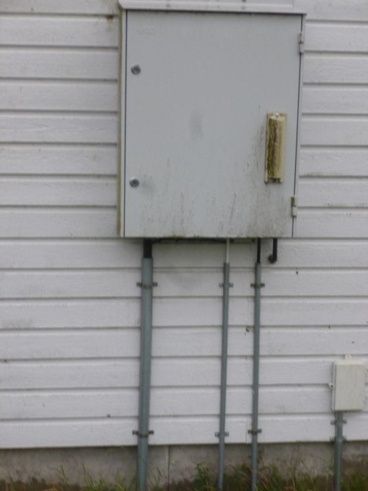 Ett fasadmätarskåp för elektricitet på en vit vägg med två kablar som leder till andra byggnader och en oansluten fiberdosa.