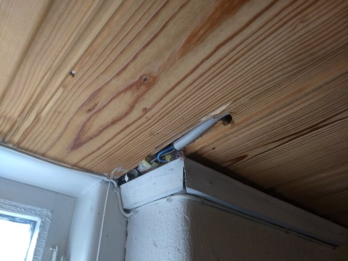 Klippt elektrisk kabel som sticker ut från ett hål i en träpanelvägg med isolering och ledningar synliga.
