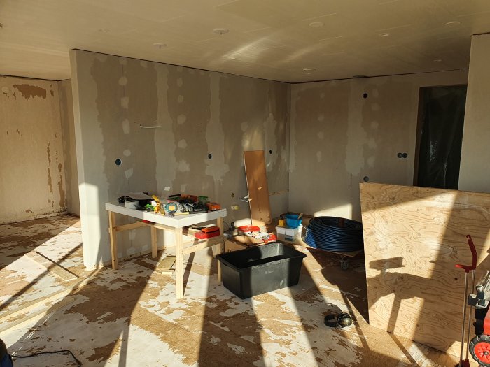 Renoveringsarbete i kök med golvvärmerör, oslipat golv och väggar klädda med plywood och gipsskivor.