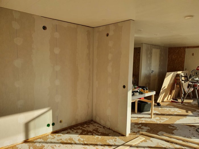 Renoveringsarbete pågår i ett rum med plywoodväggar och borttagen golvbeläggning samt byggmaterial synligt.