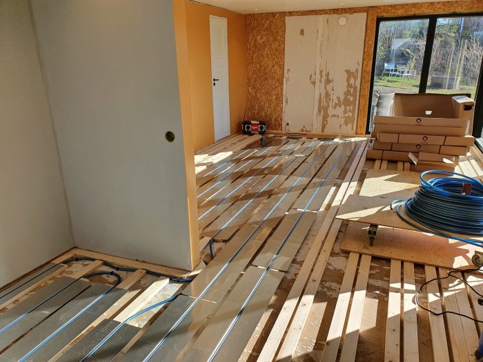 Renoveringsprojekt av kök med golvvärmerör utlagda på råspont och plywoodväggar i bakgrunden.