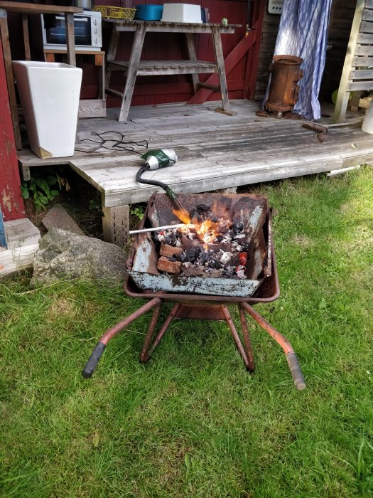 Eld i en gammal rostig skottkärra med en byggtork riktad mot lågorna, belägen på en gräsmatta.