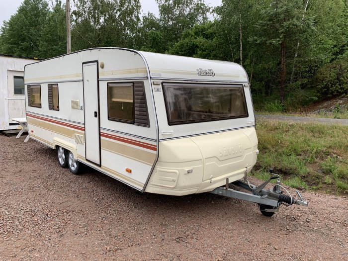 En Sävsjö 612 husvagn från 1987 parkerad på en grusyta med skog i bakgrunden.