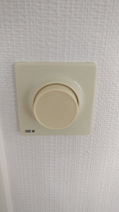 Gammal beige dimmerknapp med texten "300 W" på en vägg med vit tapet.