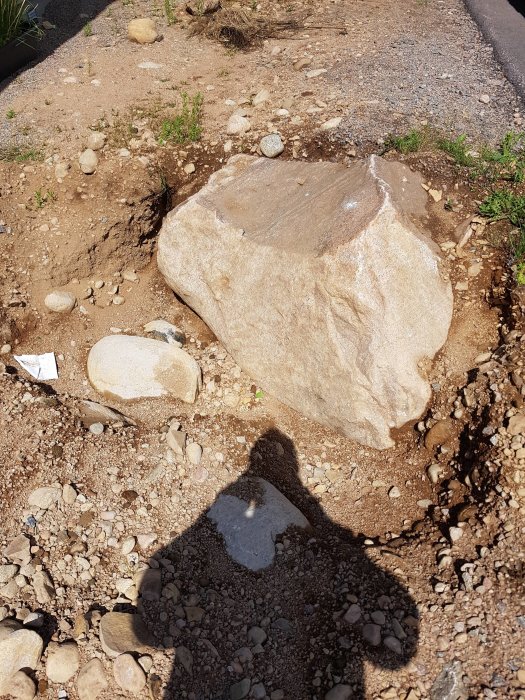 Stor sten i jordig terräng med mindre stenar runtom, lämnad efter borrning, med människoskugga i förgrunden.