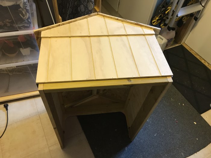 Hemmagjort småhus i trä med öppen ingång, klätt med plywood och OSB-tak, vitmålade detaljer saknas.