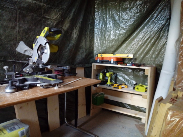Verktygshylla av trä med såg och andra verktyg i ett förrådsutrymme med plastöverdragen vägg.