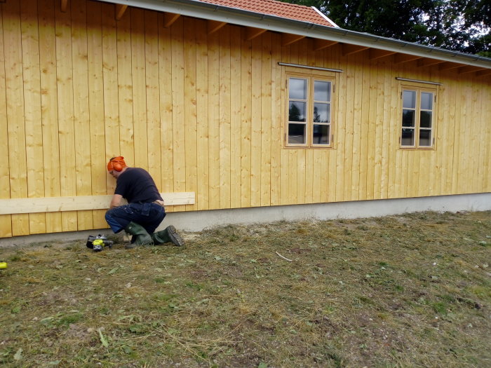 Person i arbetshjälm böjer sig ner vid ny fasad på hus, kapar bräder vid grunden.