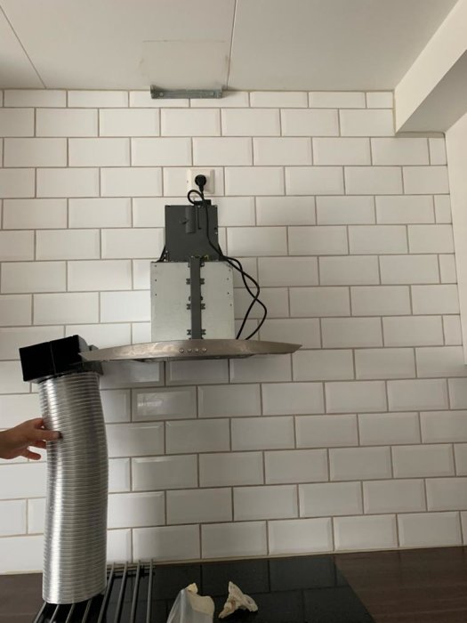 Köksfläkt monterad på vit kakelvägg utan synlig ventilation, flexibel slang hålls av en person.