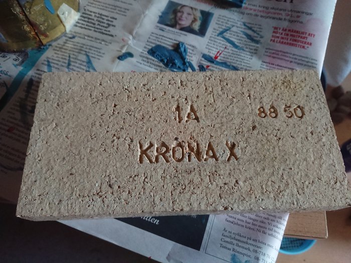 Gammal tegelsten med texten "KRÖNAX 1A 88 50" på en liggande tidning.