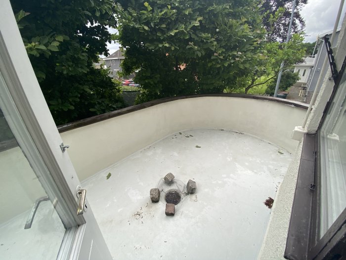 Pyöreä terassi, jossa betonilattia ja viemärikaivo. Terassin nurkassa on kiviä. Taustalla vihreitä puita ja naapuritaloja.