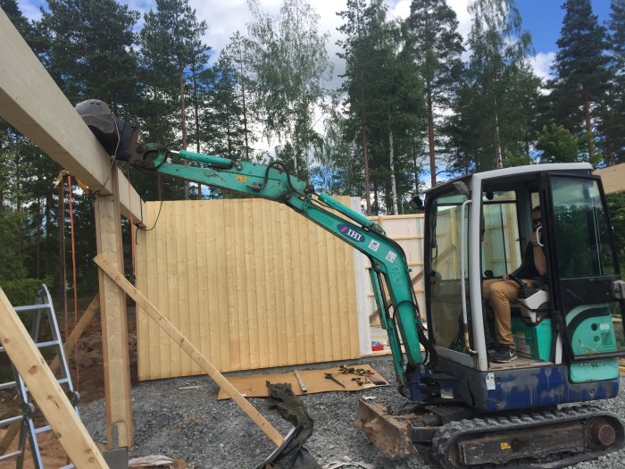 En mindre grävmaskin hjälper till att lyfta en limträbalk under konstruktionen av en carport.