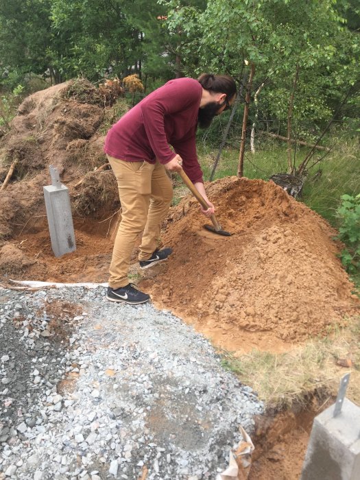 Person som gräver vid grundplint för carport, bredvid stolpsko och hög med grus.