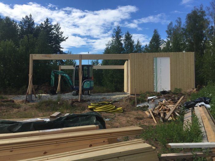 Delvis byggd carport med uppresta trästommar och grävmaskin i bakgrunden.