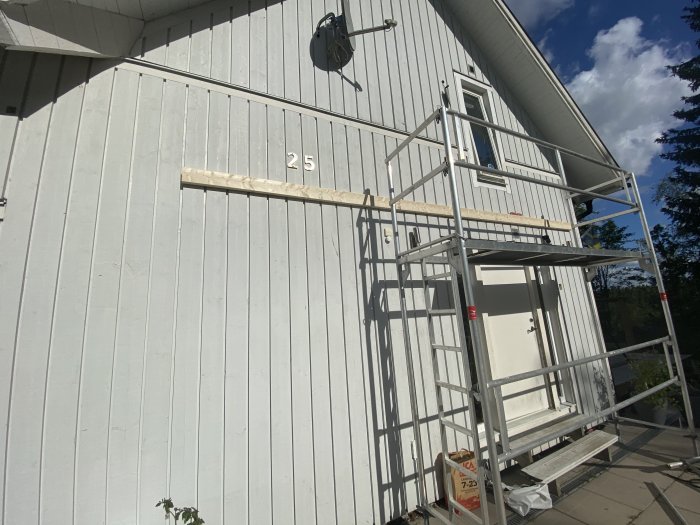 Ett hus med en vit fasad och en balk monterad vid entrén, med ställningar vid sidan, under en solig himmel.
