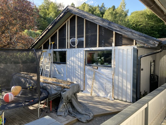 Nyuppsatt stående vit panel på ett hus med byggmaterial och stege synligt, samt en tillfällig lösning vid vattenutkastaren.