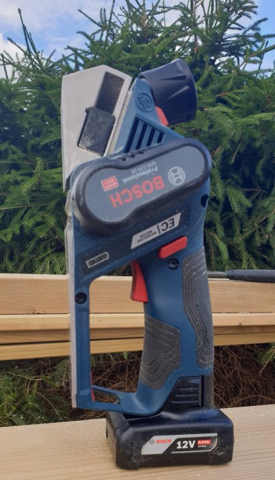 Bosch 12V elektrisk hyvel på träplanka framför granhäck.