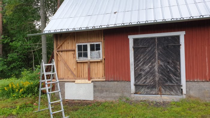 Äldre rödmålat slakteribyggnad under renovering med avlägsnad panel och synligt regelverk, bredvid en stege.