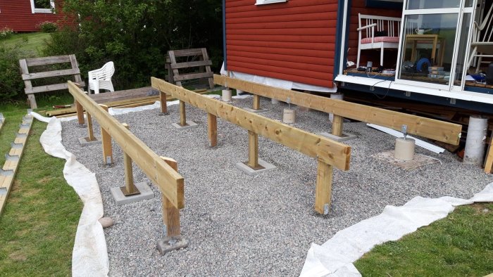 Altan under konstruktion med bärlager och stolpar på plintar, ojämn undergrund med stödmur i bakgrunden.