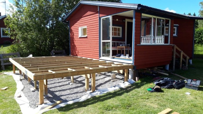 Altan under konstruktion med träreglar och plintar vid röd stuga, byggmaterial synligt på marken.