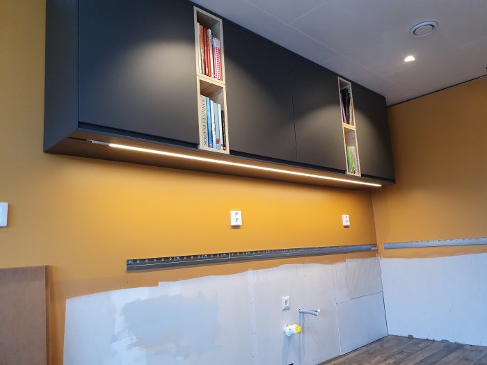 Renoverat kök med öppna IKEA överskåp och undermonterad LED-belysning, ommålad vägg och förberedelse för kakel och nya vitvaror.