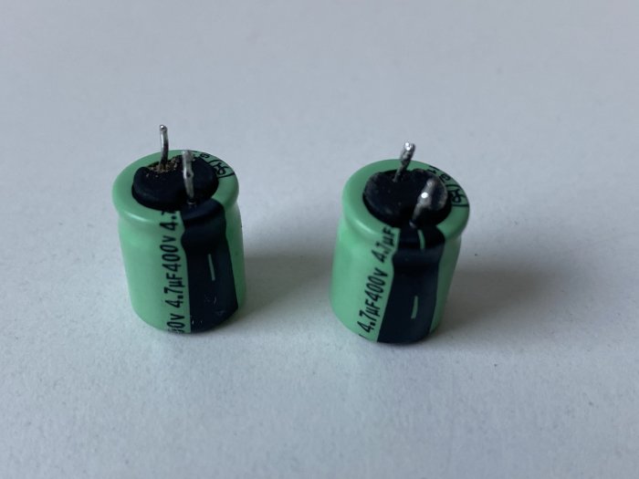Två gröna trasiga kondensatorer med svarta märken och böjda stift på vit bakgrund.