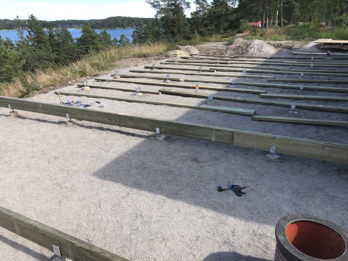 Grundläggning av altandäck med träbalkar och justerbara stolpskor på grusunderlag, vid kustmiljö.