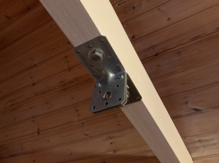 Enkel metallfästeanordning monterad på vitmålad takbjälke mot brun träpanel.
