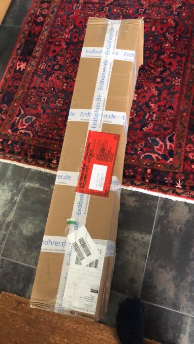Långt paket levererat till dörrmatta i en hall, med fraktetiketter på ovansidan.