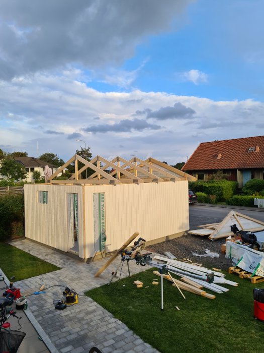 Garage under uppbyggnad med väggar uppe och takstolar som monteras, byggmaterial och verktyg syns framför.