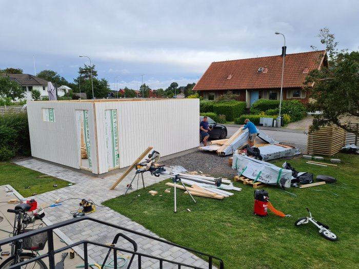 Montage av garage med personer som arbetar, byggmaterial spridda över gårdsplats.