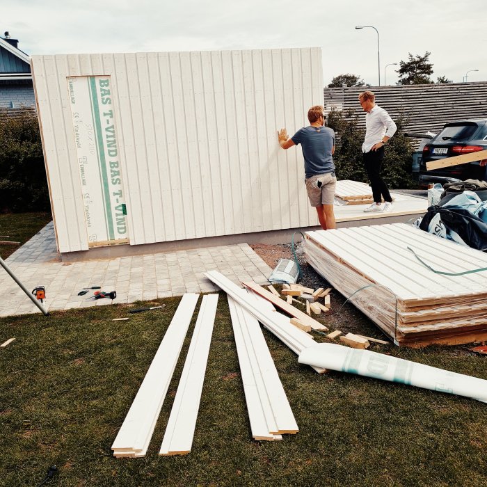 Två personer monterar en vit garagevägg, byggmaterial och verktyg syns utspridda på gräsmattan.