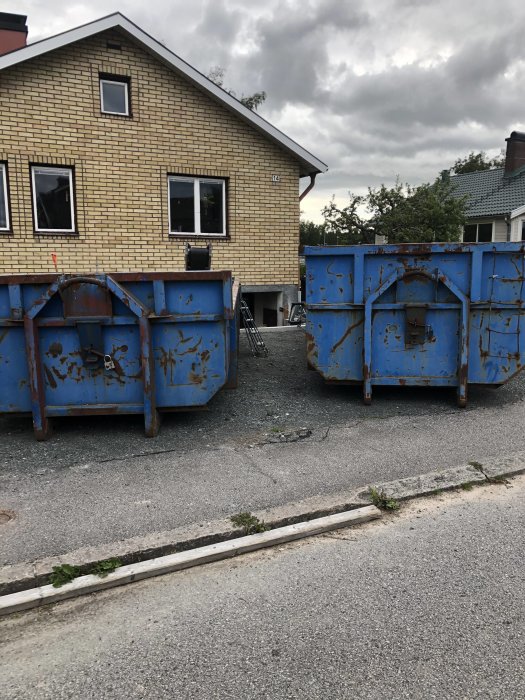 Två stora blåa byggcontainrar framför ett gulfärgat hus, indikerar pågående renoveringsarbete.