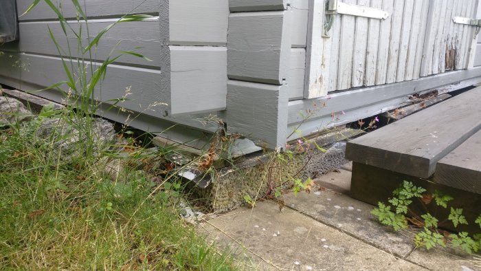 Förrådshörn med ruttnad bräda vid foten som vilar på ett lecablock, nära vegetation och trädäck.